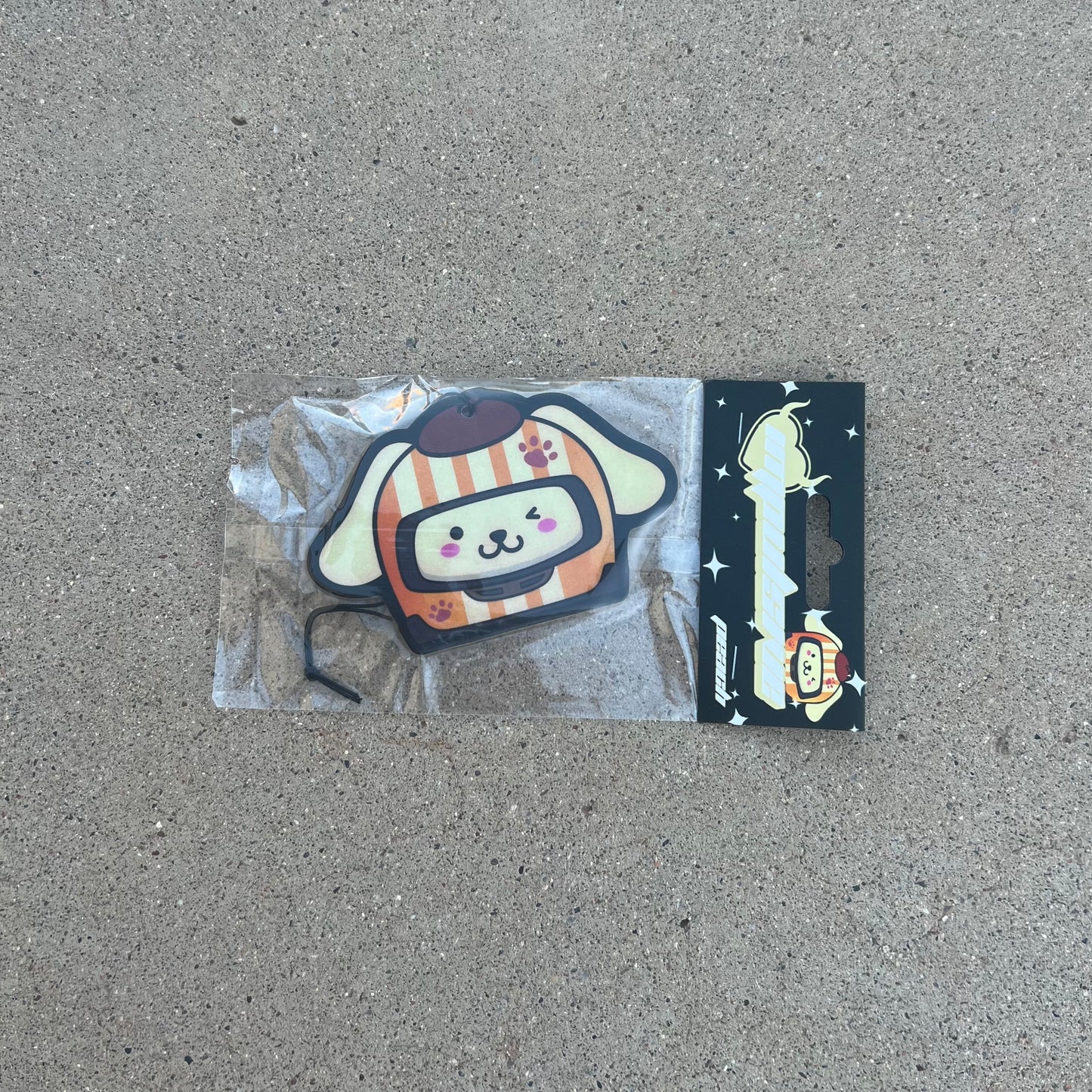 Sanrio Pompompurin Dog Racer Airfreshener hanging from black string. Cream Color dog with orange cream stripe and hat winking with paw prints on helmet. Black Background with stars and impulsive cream color y2k logo with peach label cardboard head card packaging.