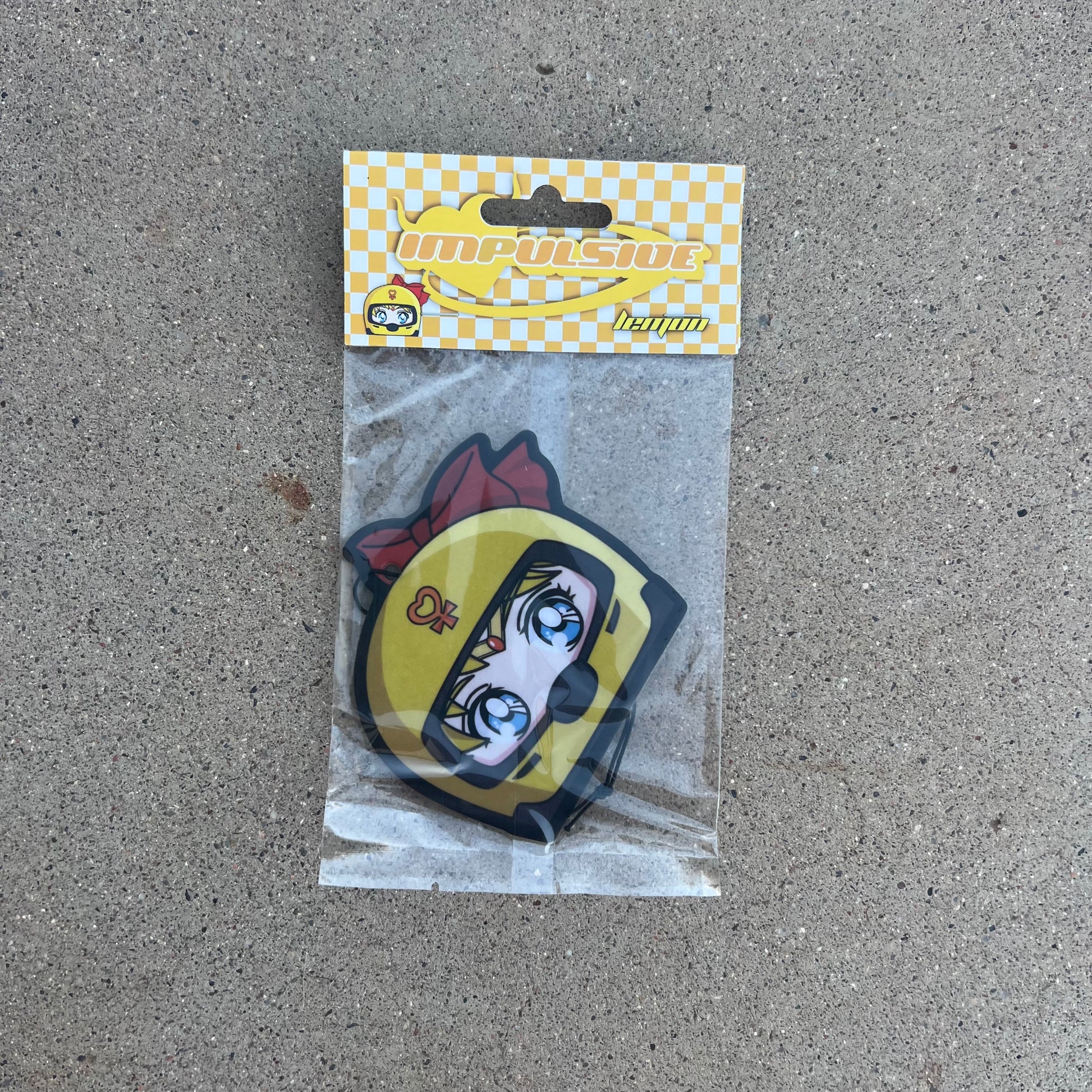 Sailor Venus air freshener hanging from black string. Japanese anime character blonde hair blue eyes wearing yellow helmet with pendent logo and red bow. yellow and white checkered background with yellow y2k impulsive logo and lemon scent label cardboard head card packaging.