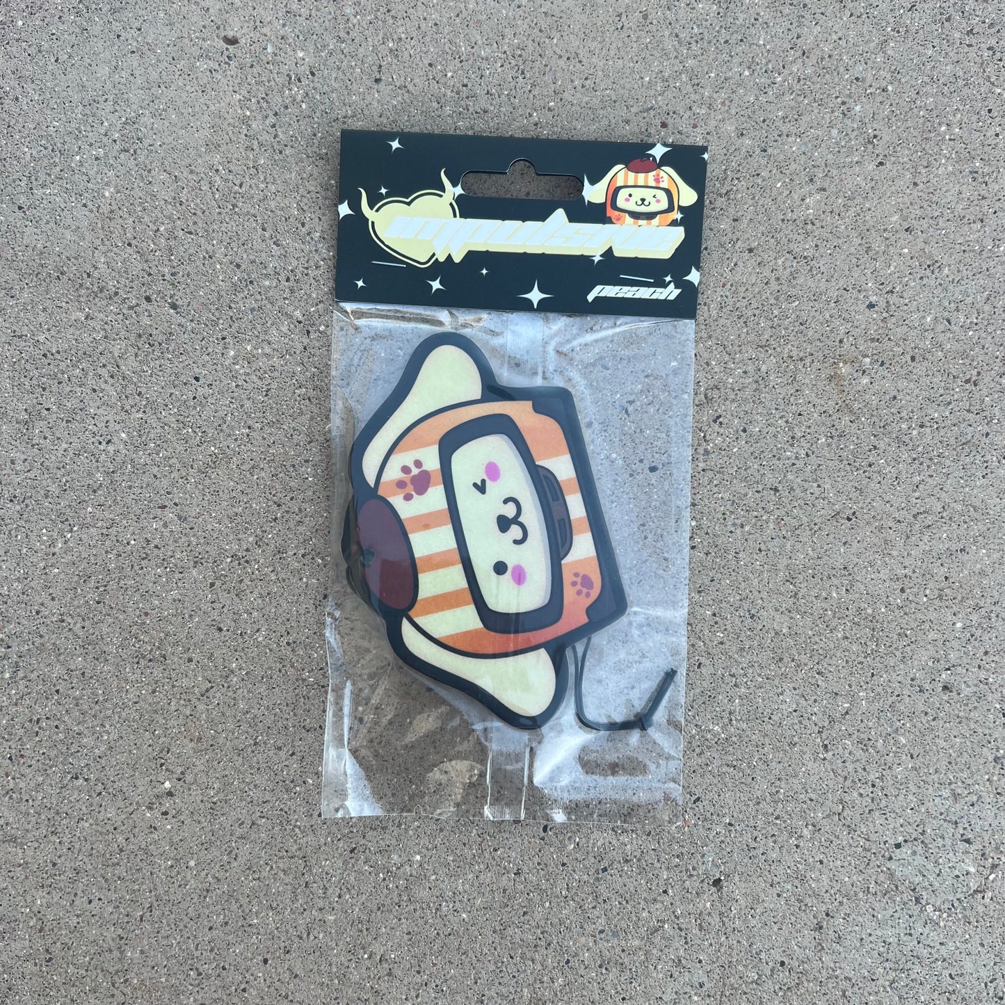 Sanrio Pompompurin Dog Racer Airfreshener hanging from black string. Cream Color dog with orange cream stripe and hat winking with paw prints on helmet. Black Background with stars and impulsive cream color y2k logo with peach label cardboard head card packaging.