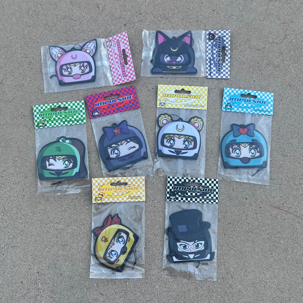 Multiple Anime Designs hanging from black string air freshener pack. Sailor moon in white helmet. Sailor Venus in yellow helmet. Sailor Mercury in blue helmet. Sailor Mars in purple helmet winking. Chibi USA in pink helmet. Tuxedo Mask in black helmet with tall hat. Luna Cat in gray Helmet with cat ears.  Color Coded Checkered Cardboard packaging 
