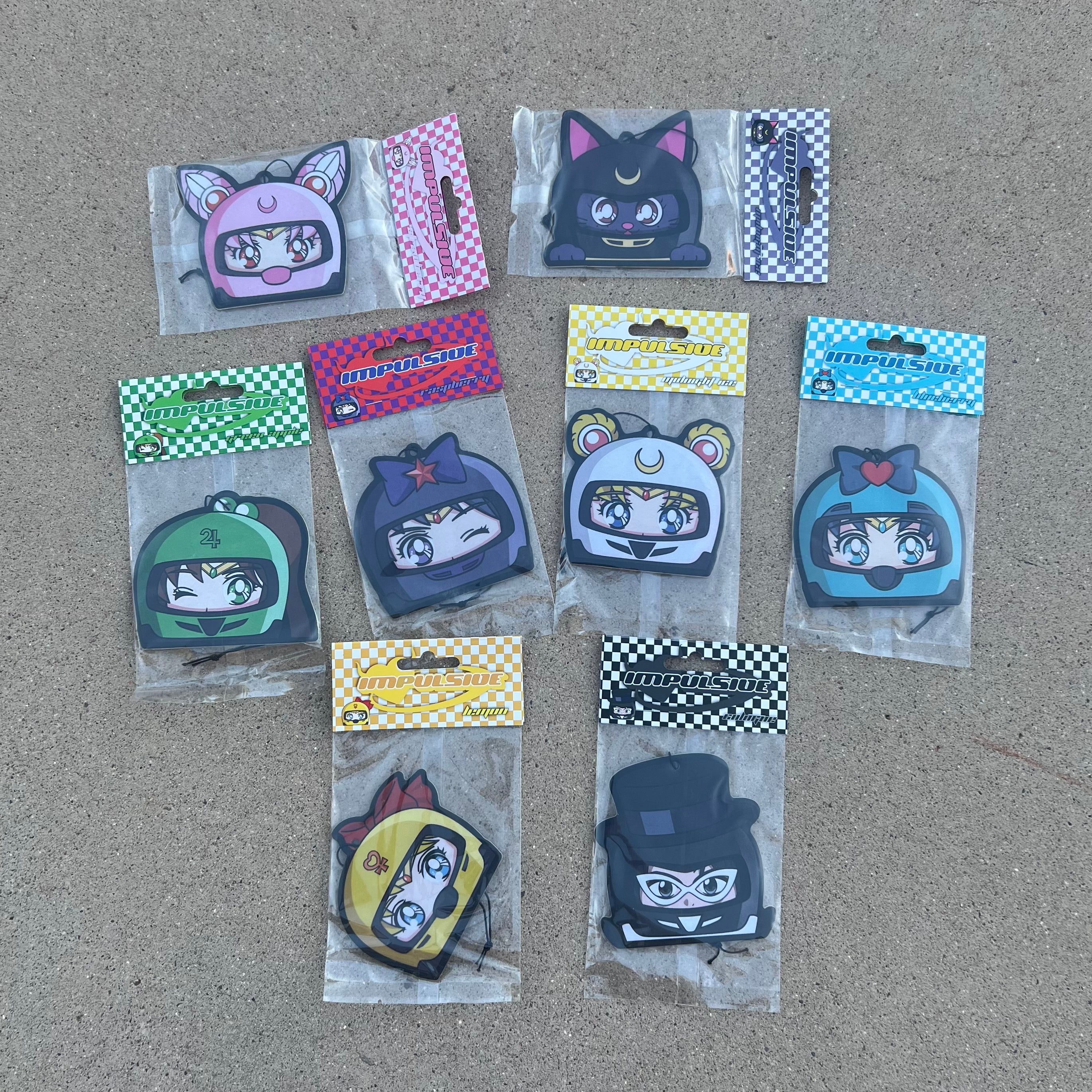 Multiple Anime Designs hanging from black string air freshener pack. Sailor moon in white helmet. Sailor Venus in yellow helmet. Sailor Mercury in blue helmet. Sailor Mars in purple helmet winking. Chibi USA in pink helmet. Tuxedo Mask in black helmet with tall hat. Luna Cat in gray Helmet with cat ears.  Color Coded Checkered Cardboard packaging 