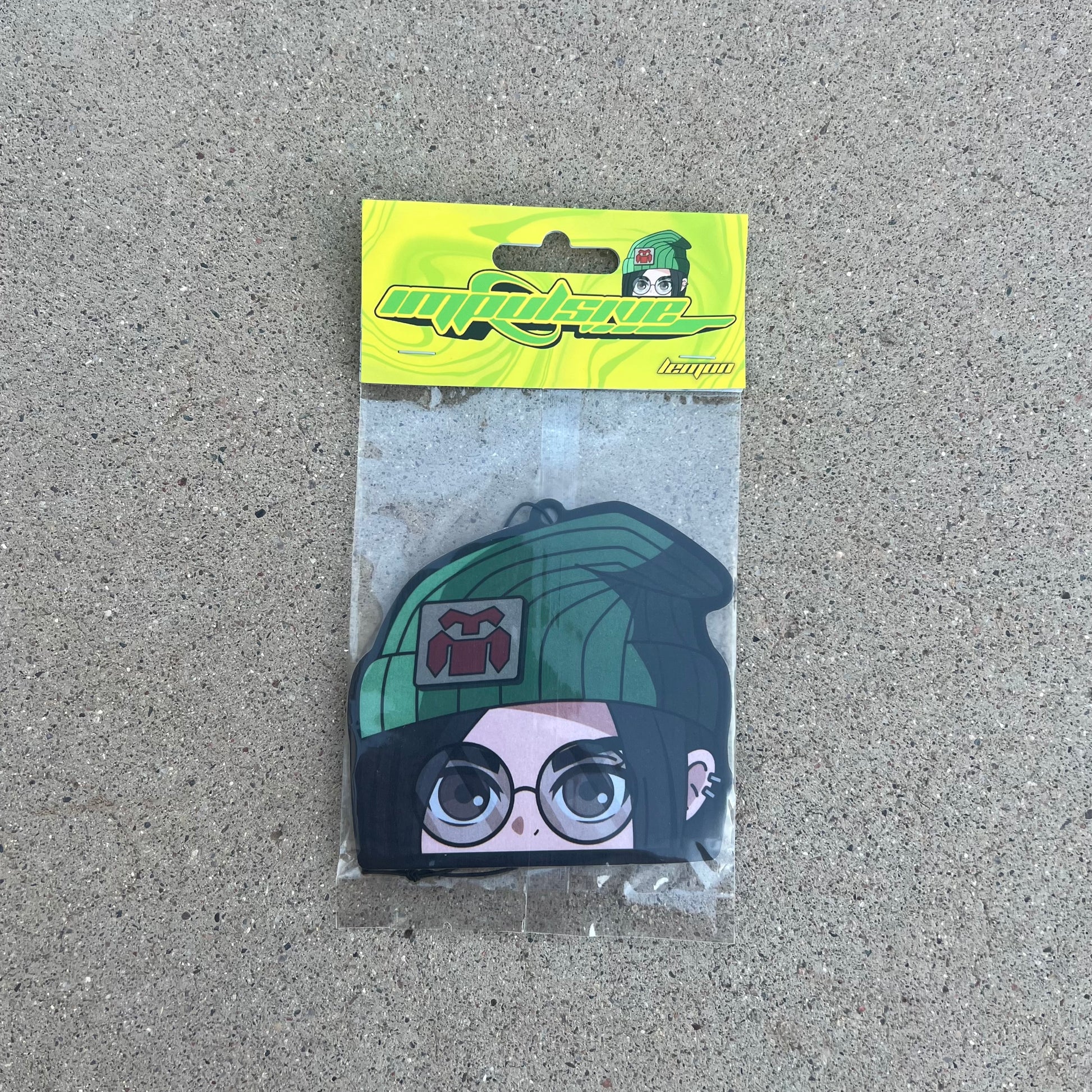 Anime Character Killjoy Lemon Scent Air freshener. Anime character, green beanie with logo, brown eyes with glasses, black hair with cuff earrings on left ear, hanging from black string. Yellow and green swirl cardboard head card packaging. Retro Y2K impulsive logo lemon label.