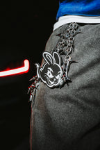A close-up of a person wearing gray textured pants and a blue top, with a heavy silver chain belt featuring a black-and-white bunny emblem and spiked hardware.