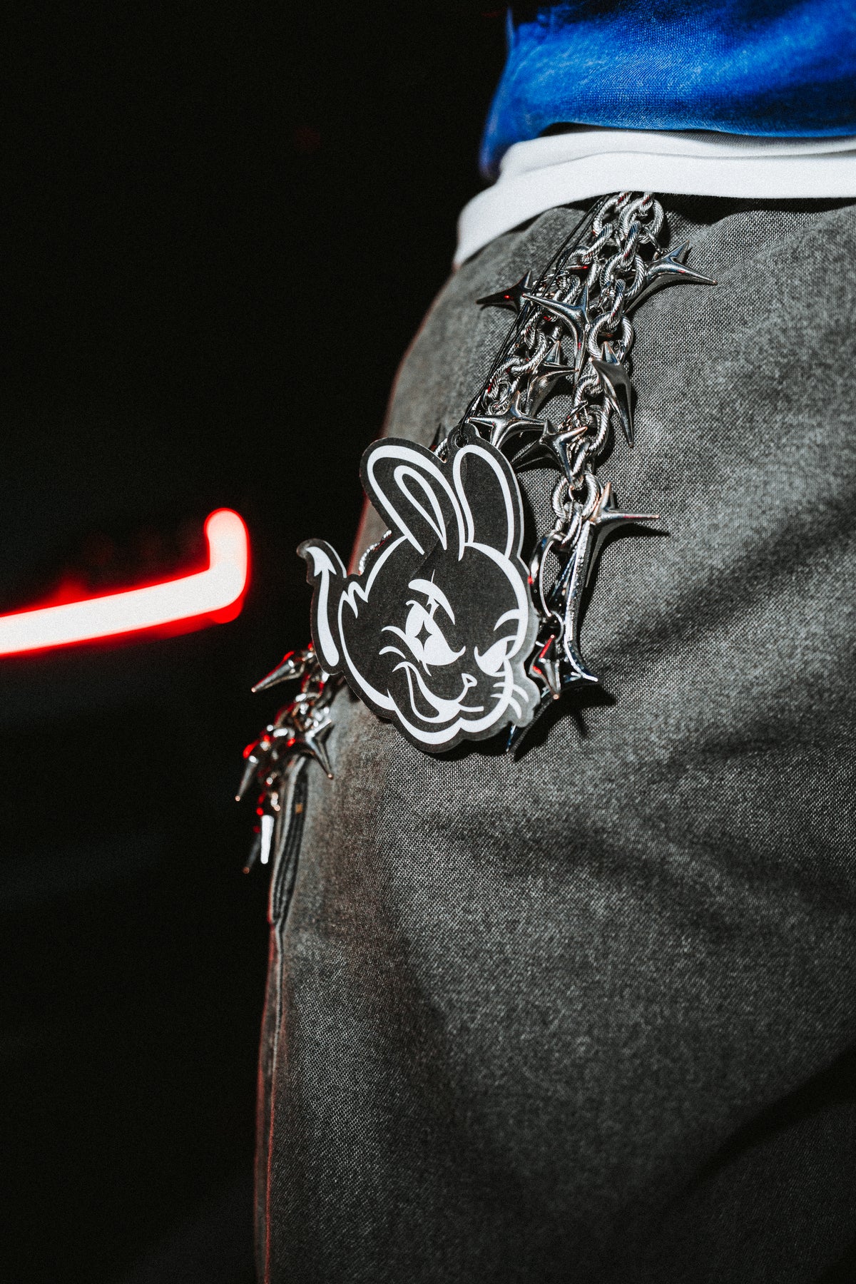 A close-up of a person wearing gray textured pants and a blue top, with a heavy silver chain belt featuring a black-and-white bunny emblem and spiked hardware.