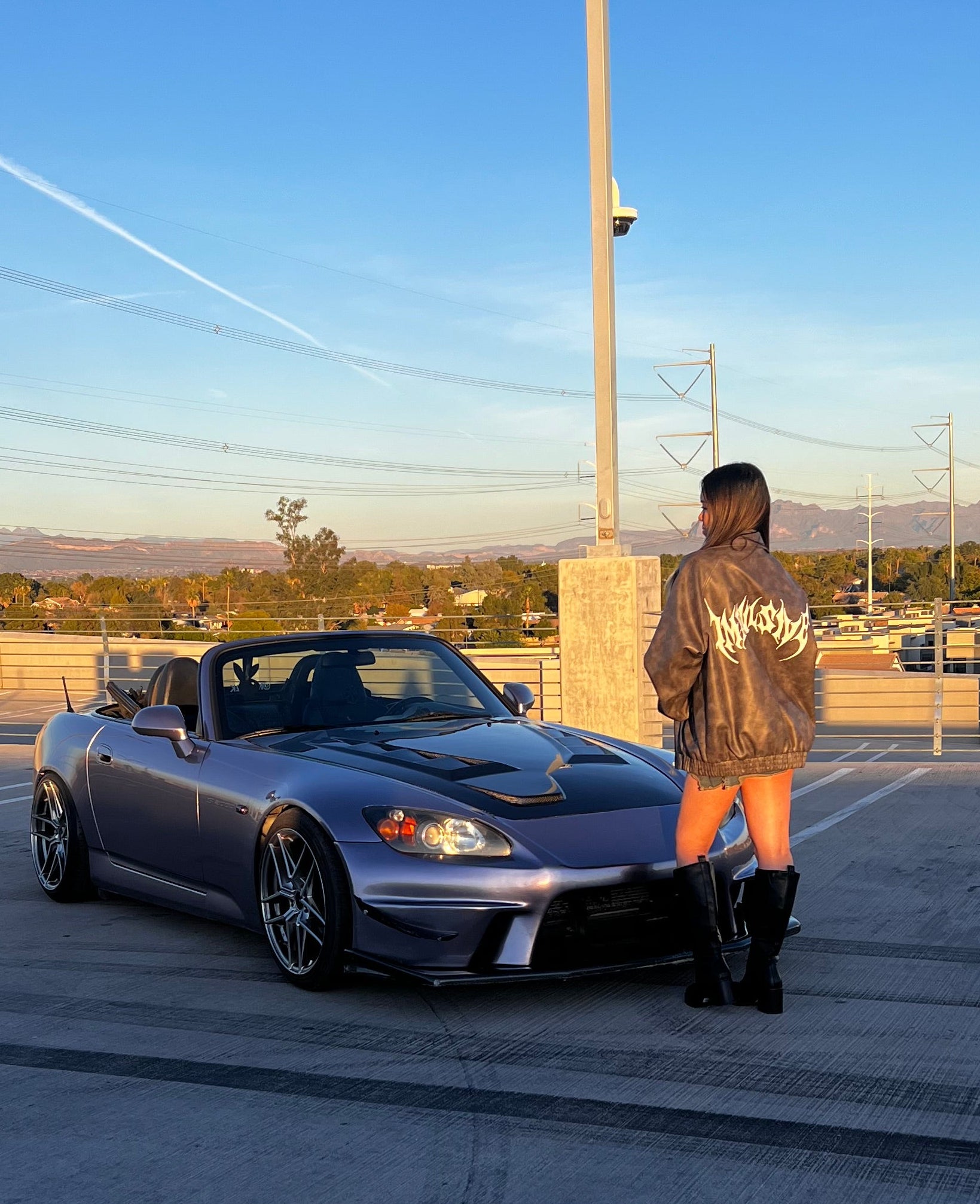 Person standing next to a silver sports car on a road with a clear sky.