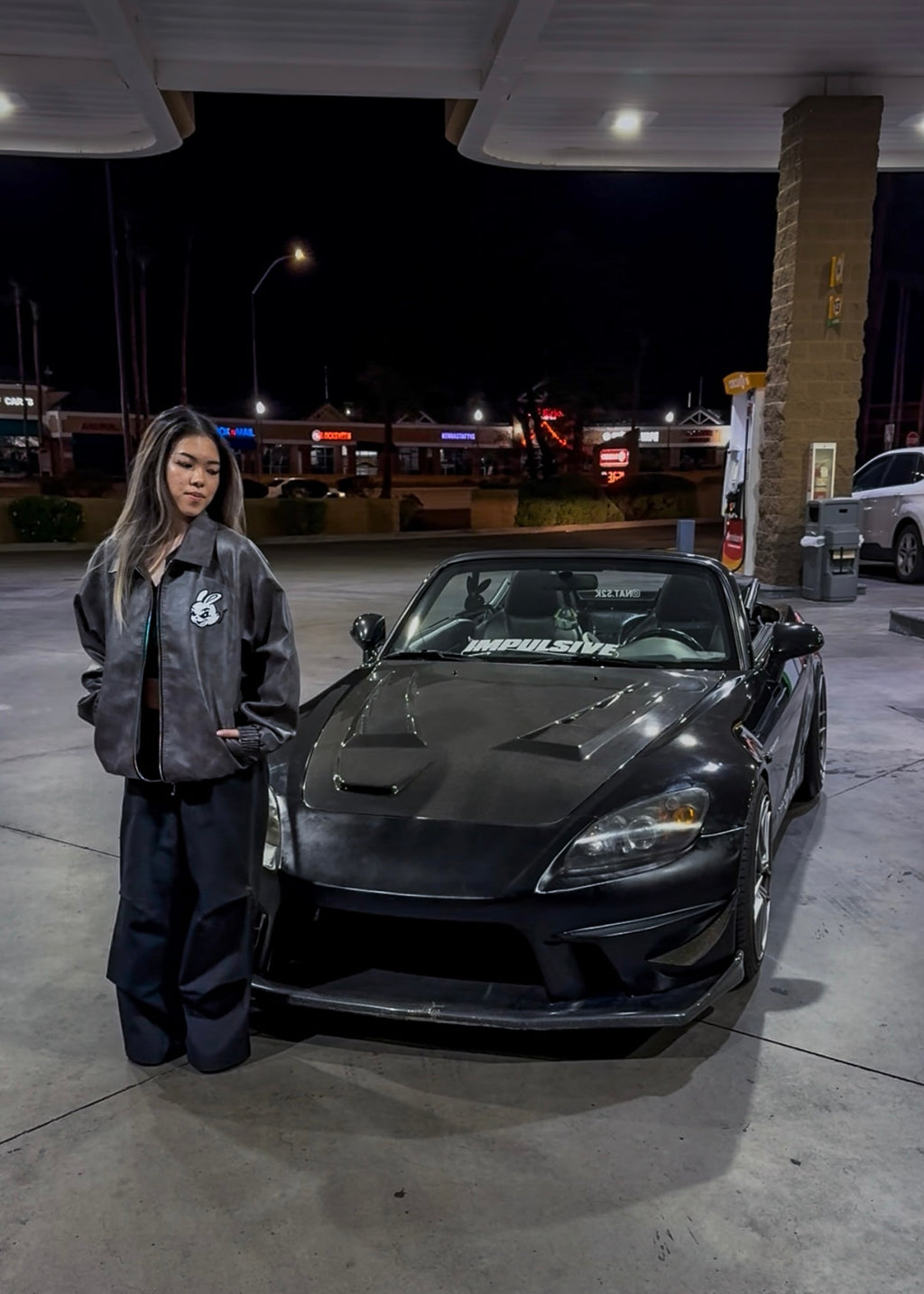 Person standing next to a black sports car honda s2000 at a gas station at night.