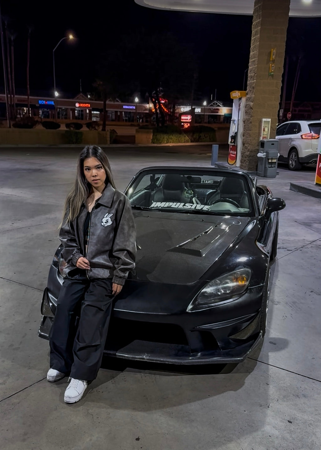 Person standing next to a black sports car at a gas station at night.